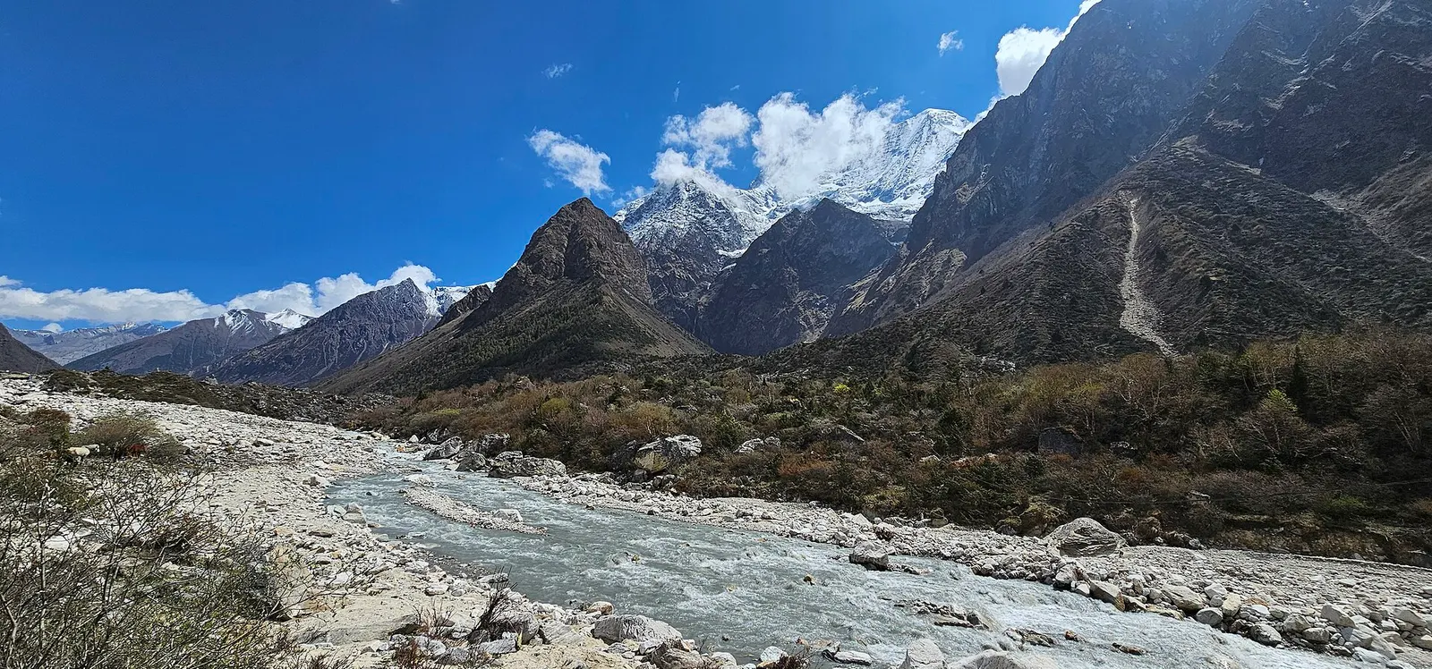 A stony glacial riverbed near Samagaon with dark slopes and snow peaks of the Manaslu range behind.