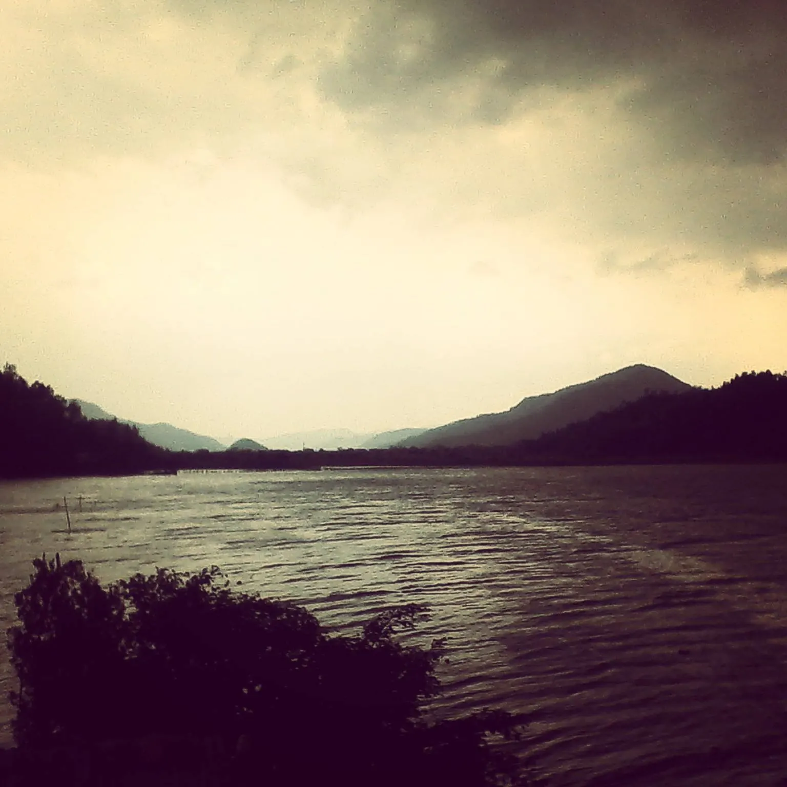 Rupa Lake under a hazy evening sky, framed by low wooded ridges.