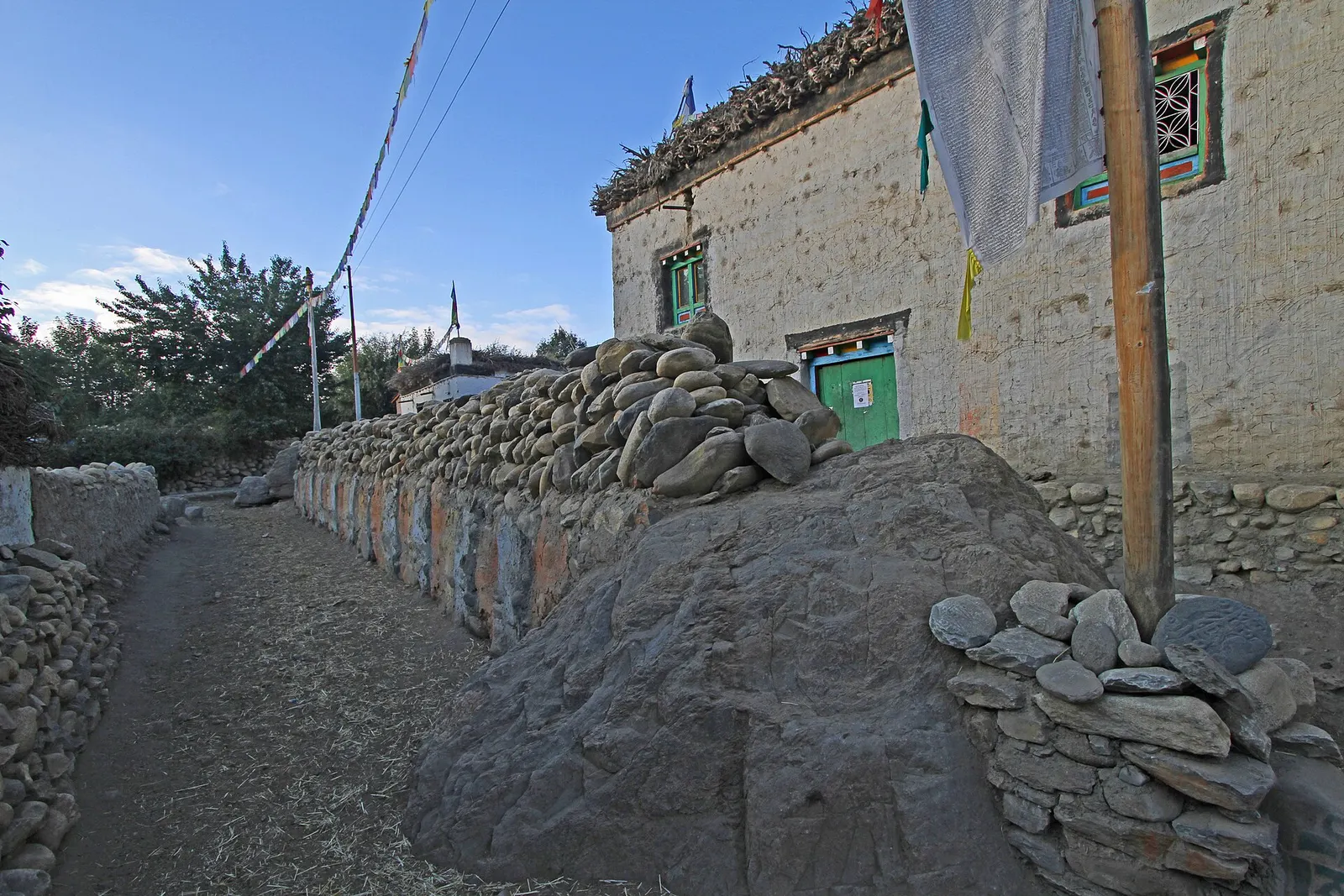 A lane in Ghami running beside a long mani wall of stacked prayer stones and a whitewashed house with prayer flags.