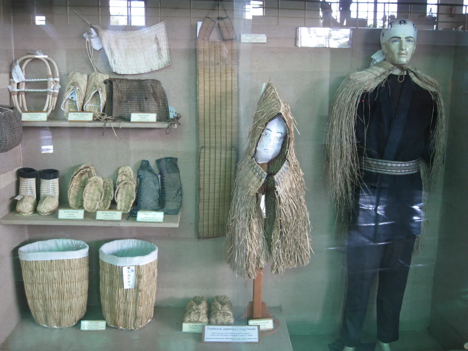 A glass display case with a straw rain cape, woven baskets, footwear and traditional clothing of Himalayan hill communities.