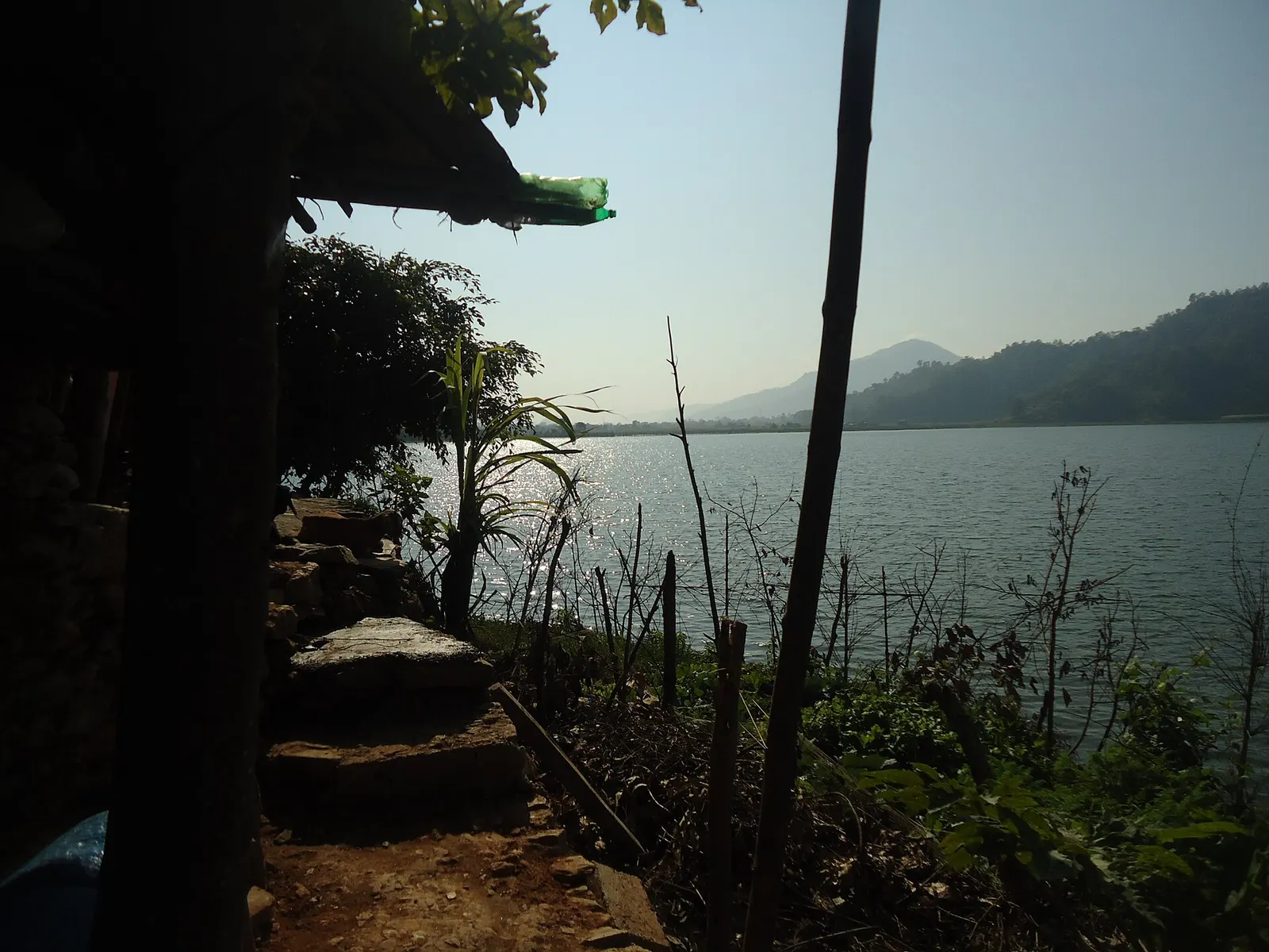 Rupa Lake seen through trees from a path on the shore, with forested hills beyond.