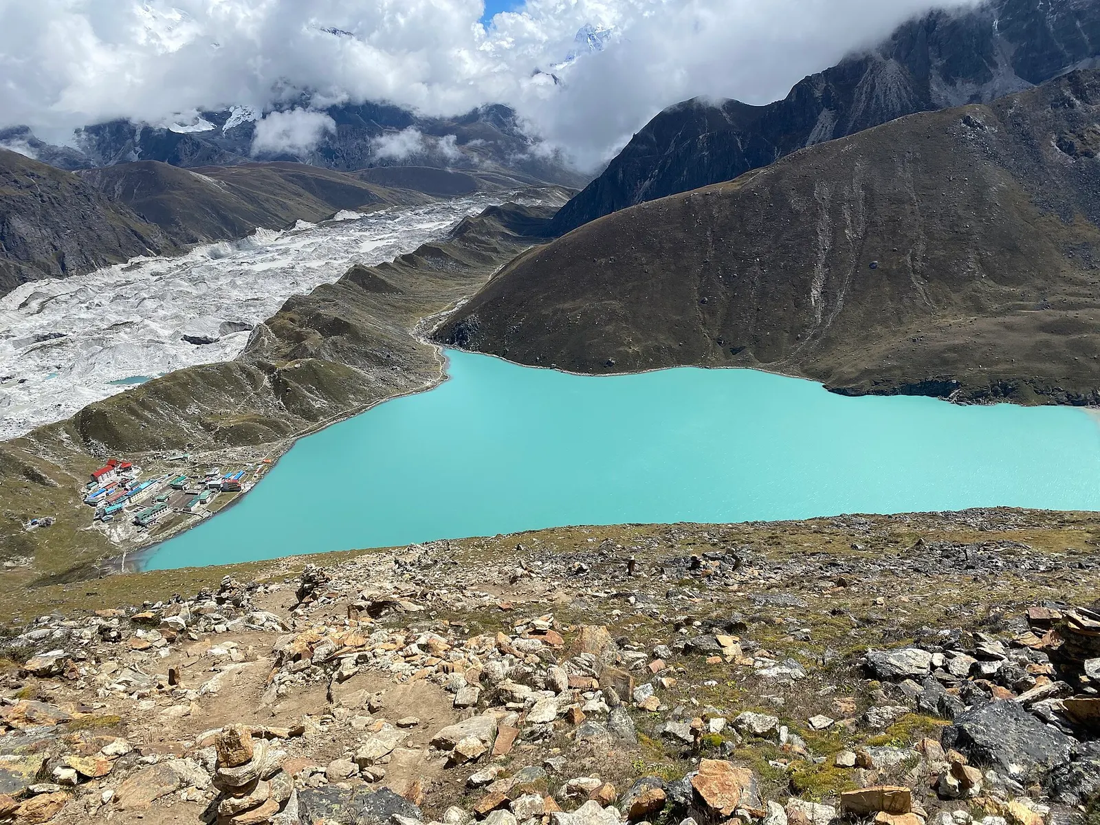 The grey rubble of the Ngozumpa Glacier beside the lodges of Gokyo village and the turquoise third Gokyo lake, seen from above.