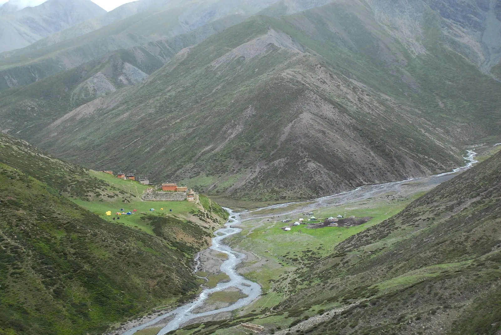 Green valley floor with a winding stream and the buildings of Shey Gompa beneath steep grassy mountainsides in Upper Dolpo.